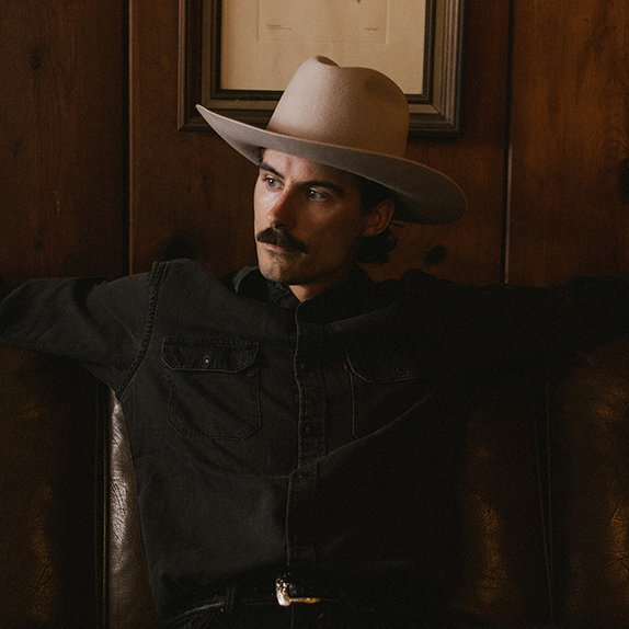 Man wearing a cowboy hat and dark shirt in a dimly lit room.