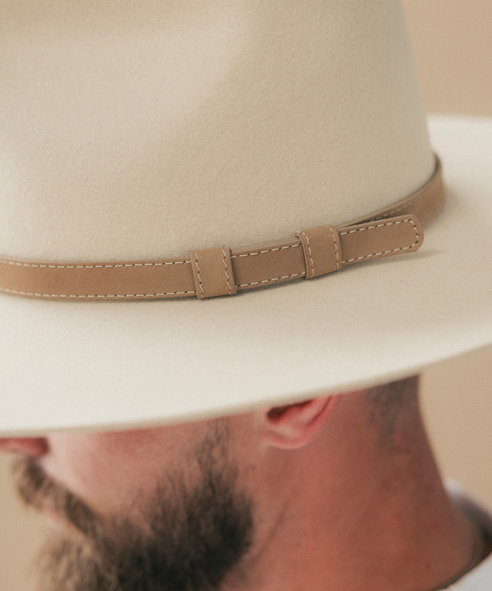 A close-up of a man wearing a cream-colored felt fedora hat with a tan leather band, with his beard and lower face visible below the brim #color_tan