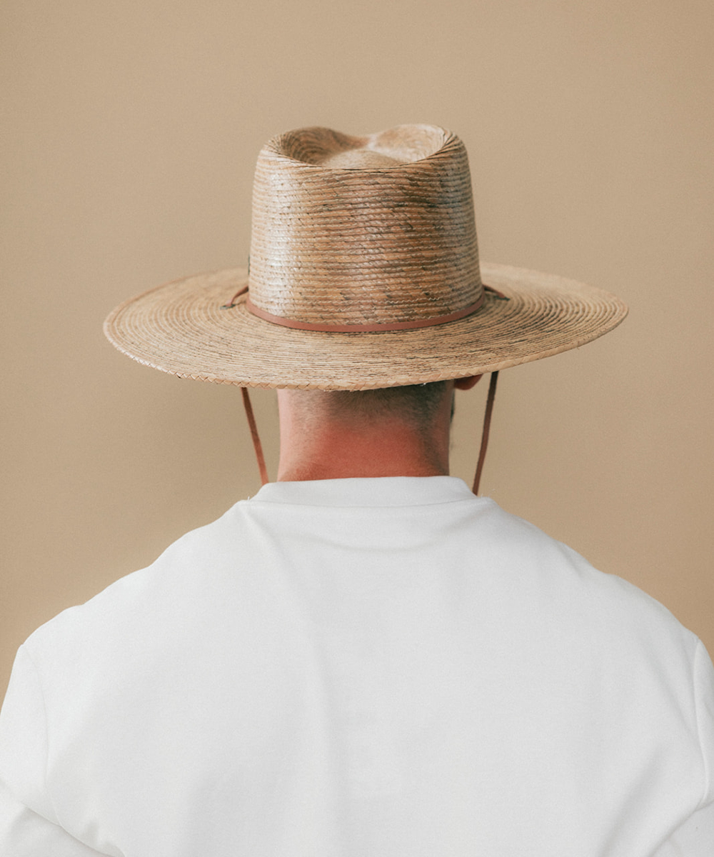 Back profile of a man wearing a natural straw cowboy hat with a brown leather band and a small silver-toned decorative emblem, against a plain beige background #color_toasted