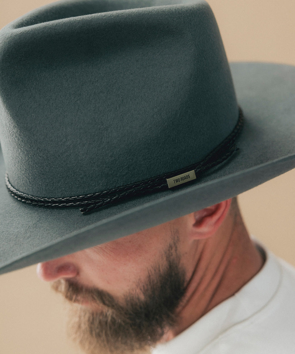 A close-up of a man wearing a dark slate gray felt cowboy hat with a black braided band and a small gold-tone metal brand tag that reads 'TWO ROADS' #color_black