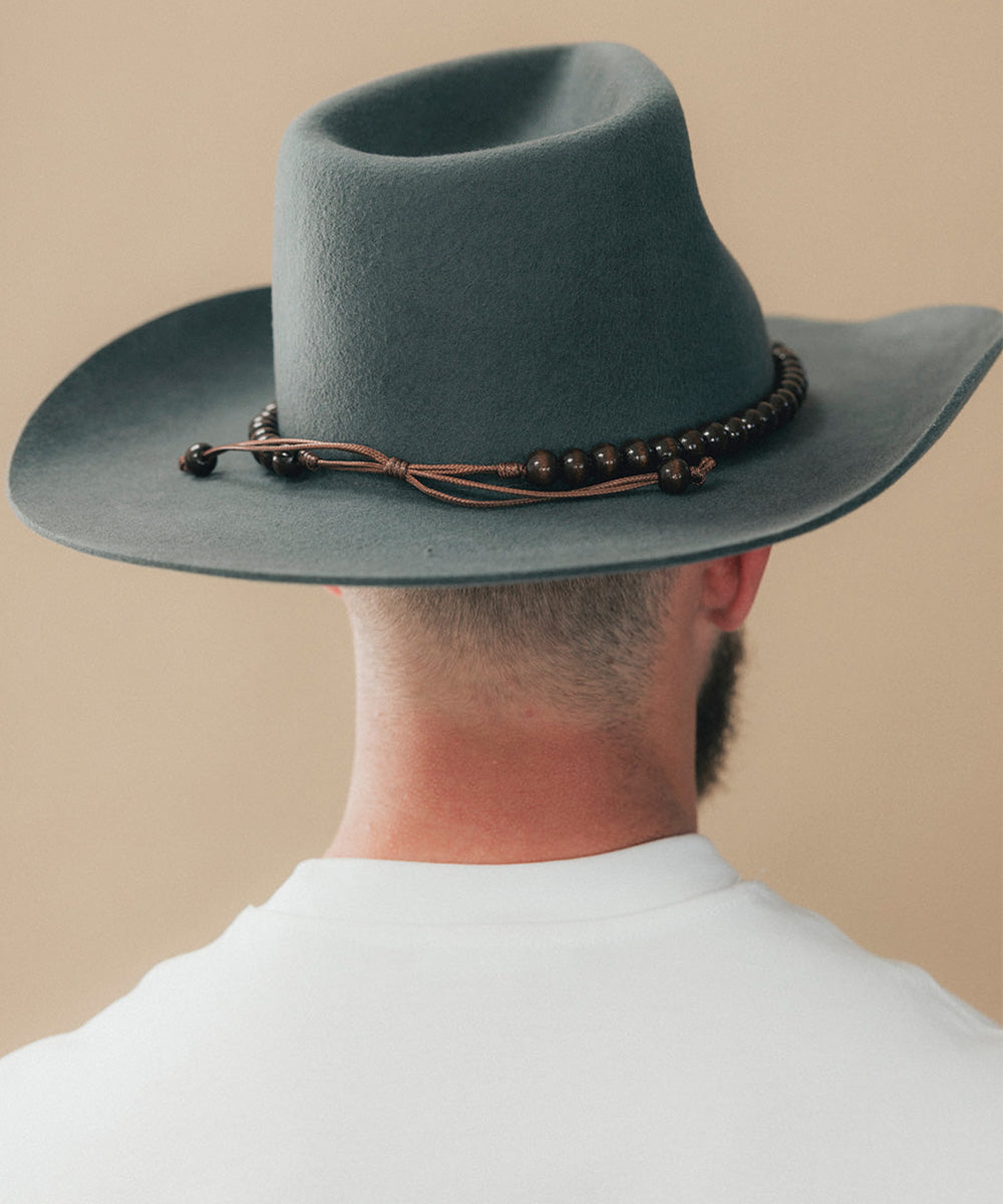 A man viewed from behind wearing a wide-brimmed felt cowboy hat in a muted blue-gray color, accented with a dark brown beaded band and cord, against a plain beige background #color_brown