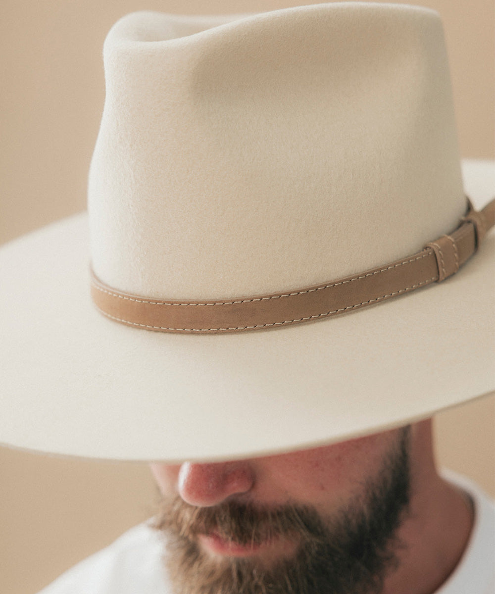A close-up of a man wearing a cream-colored felt fedora hat with a tan leather band, with his beard and lower face visible below the brim #color_tan