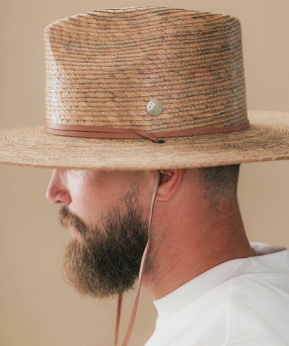 A close-up side profile of a man wearing a natural straw cowboy hat with a brown leather band and a small silver-toned decorative emblem, against a plain beige background #color_toasted