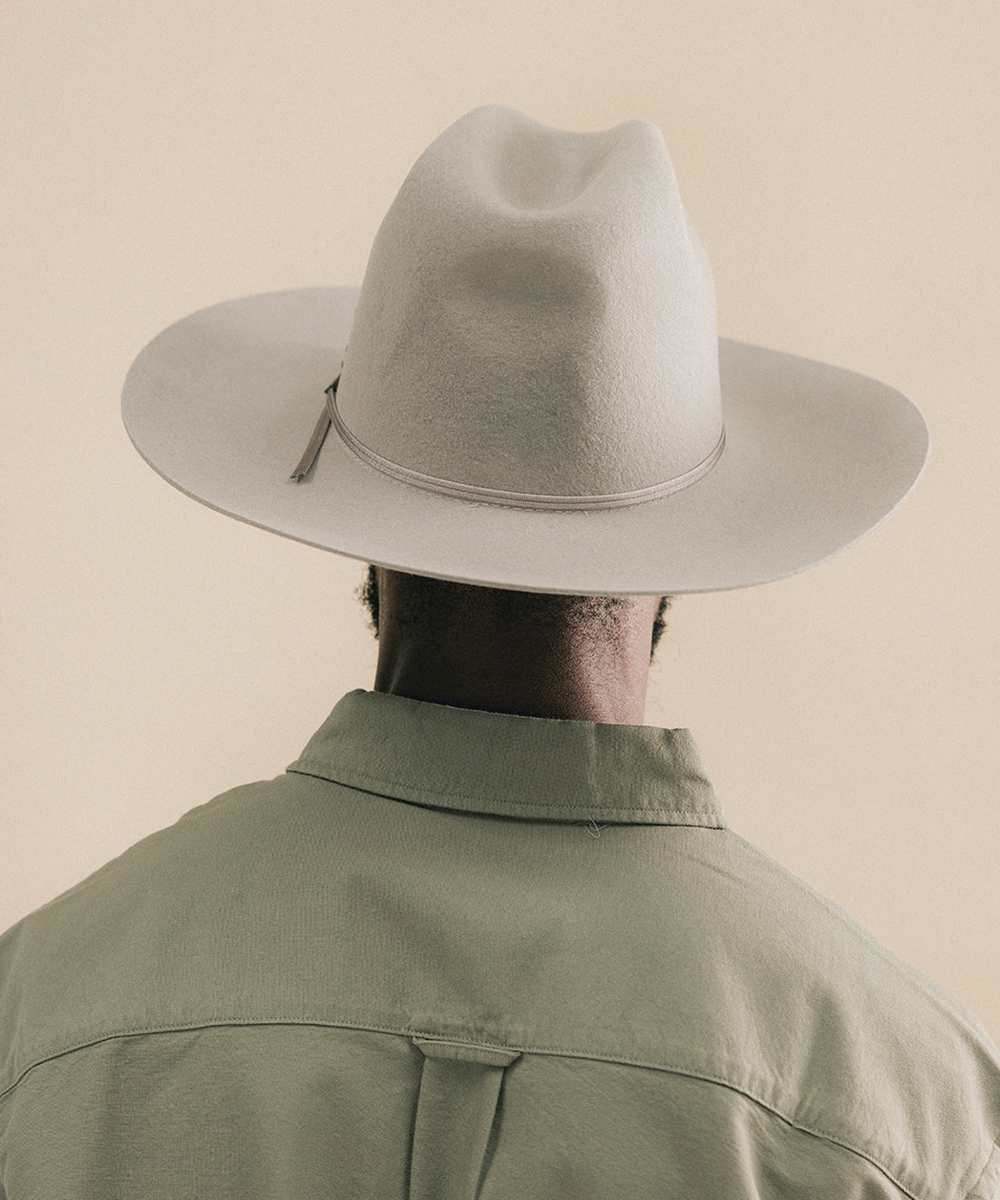 A person shown from behind wearing a light beige wide-brim fedora hat and an olive green shirt against a solid beige background #color_stone