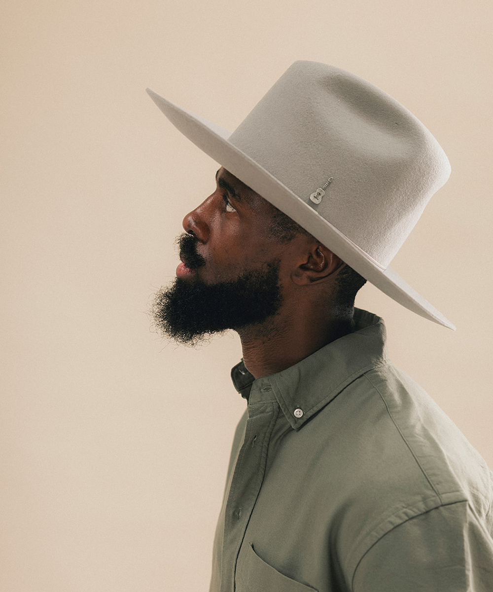 A man in profile wearing a light beige wide-brim fedora hat and a muted sage green button-down shirt, set against a solid warm beige background #color_stone