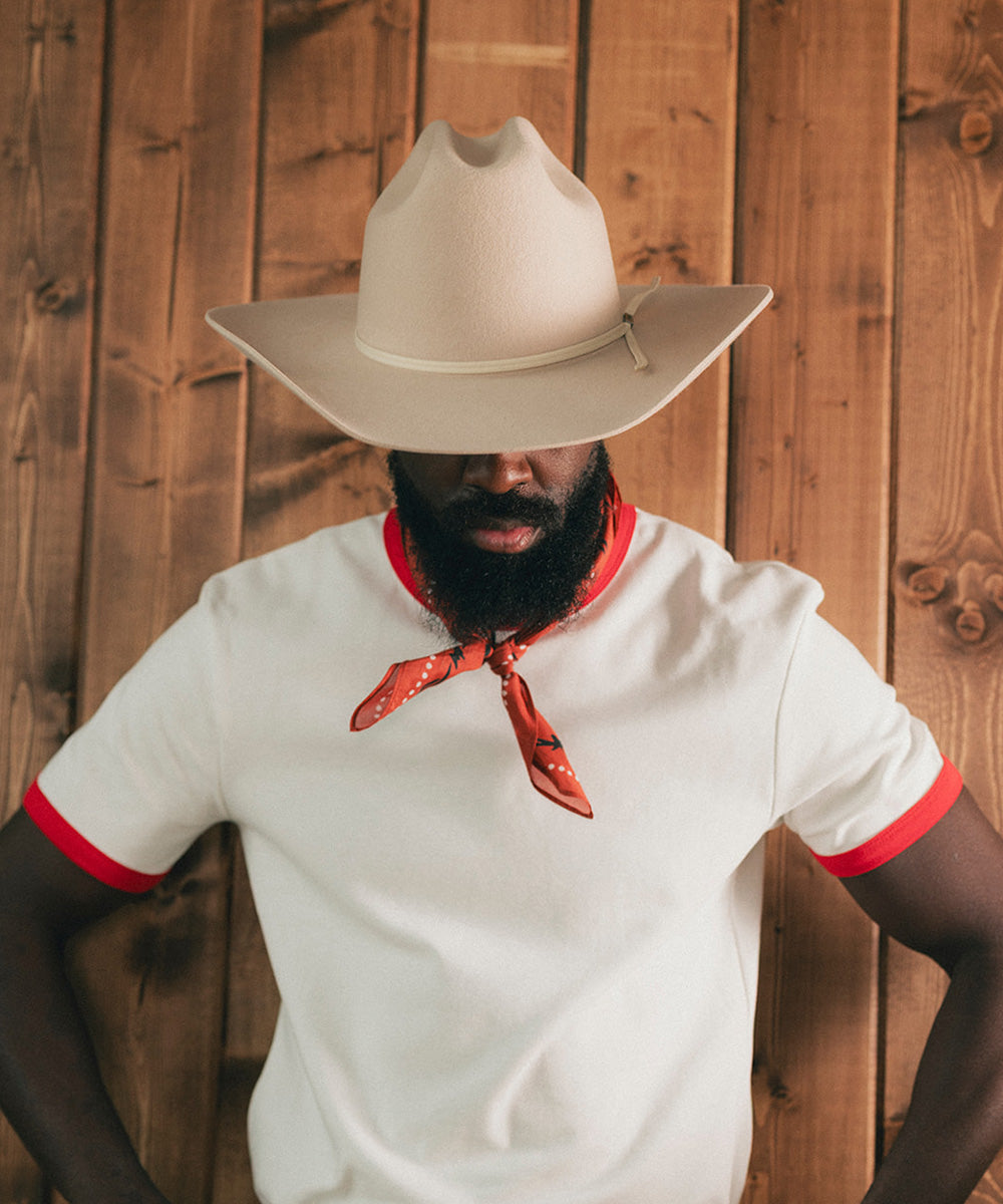Man wearing a white shirt with red trim and a beige cowboy hat against a wooden background #color_buckskin