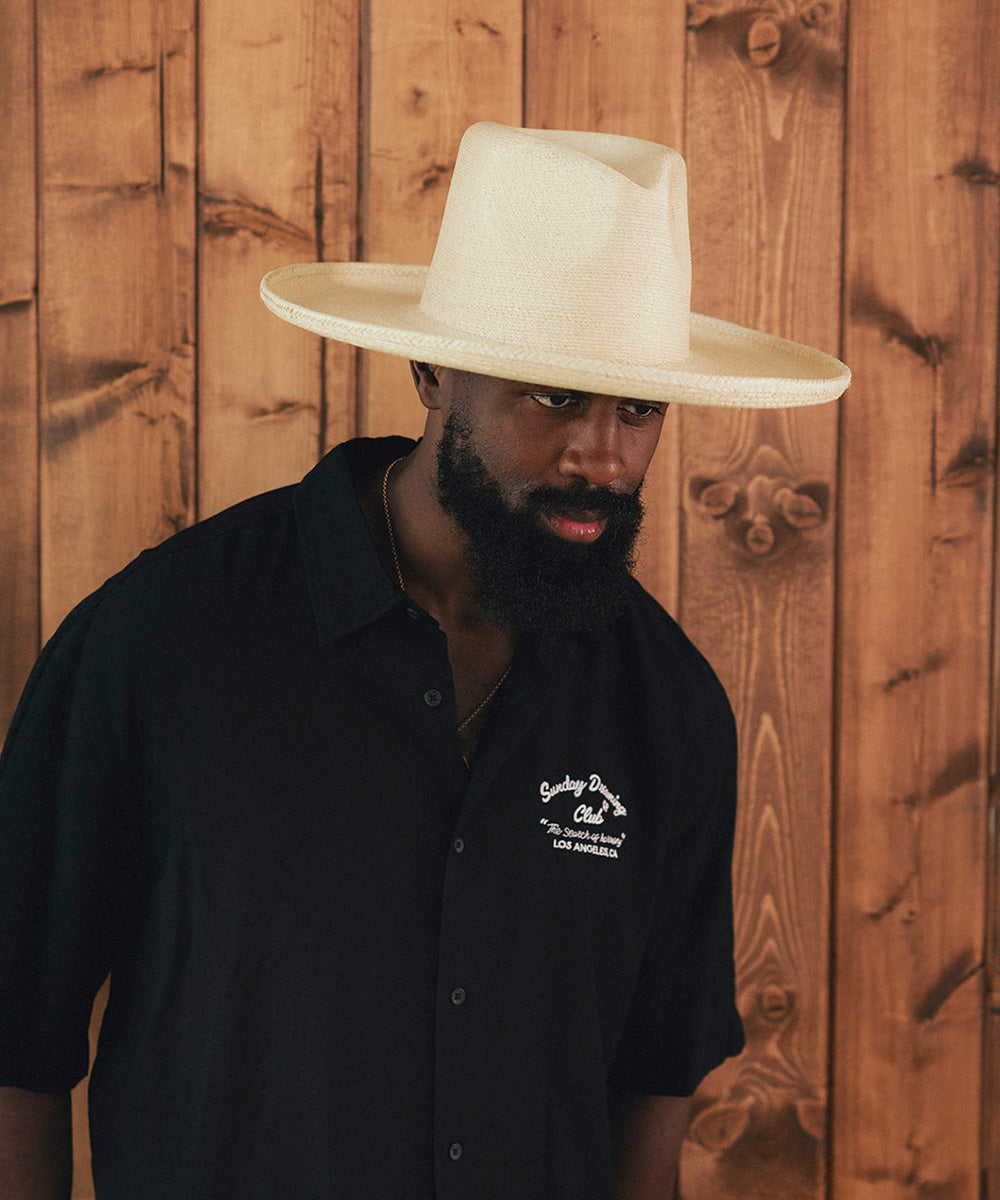 Man wearing a wide-brimmed hat and black shirt with a logo against a wooden background #color_ivory