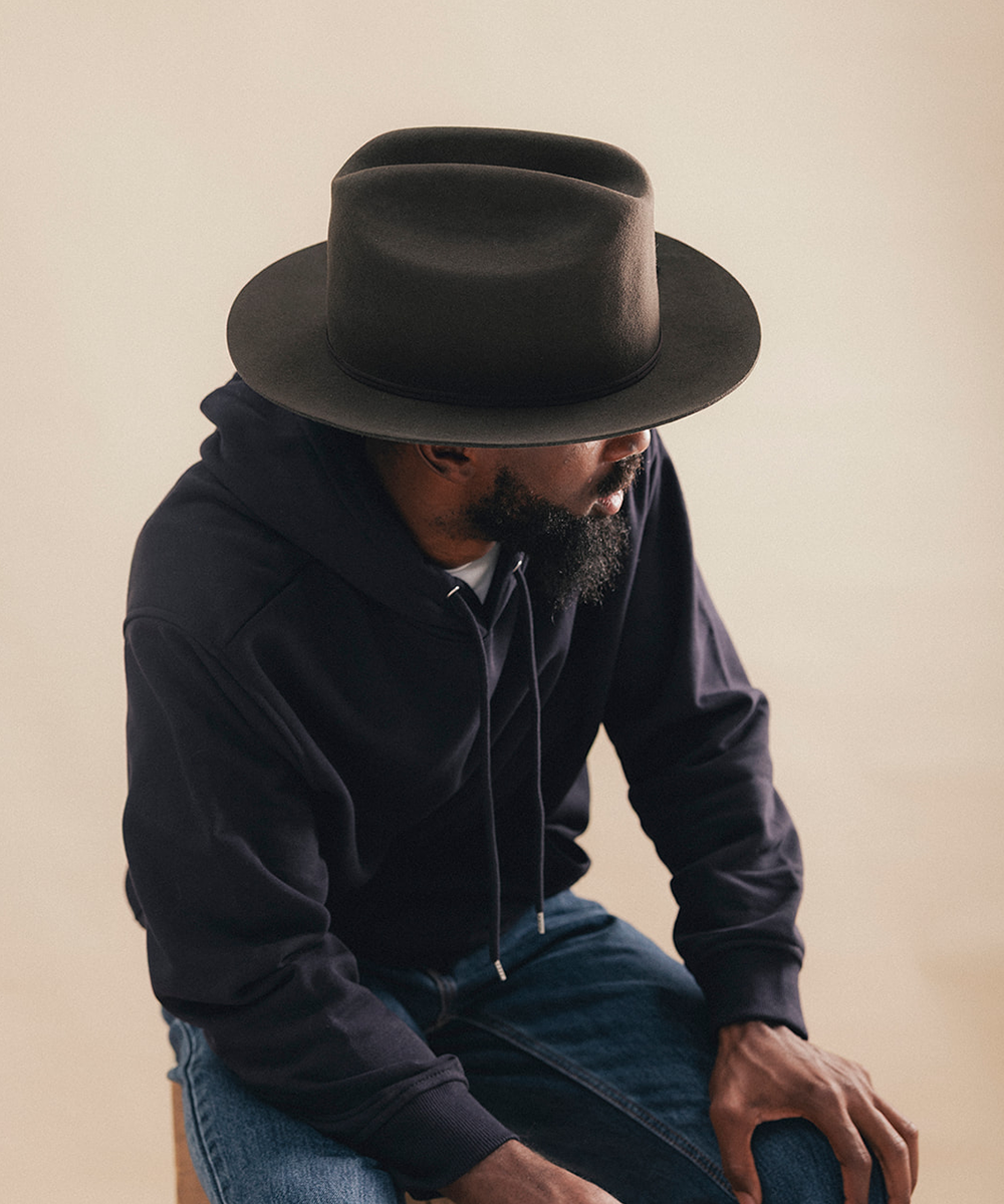 A man wearing a dark brown wide-brim fedora hat, a black hoodie, and blue jeans, sitting against a plain beige background #color_hickory