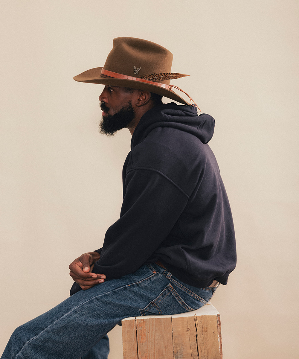 A man in profile wearing a brown cowboy hat with a tan band, a dark navy hoodie, and blue jeans, sitting on a wooden block against a plain beige background #color_tobacco