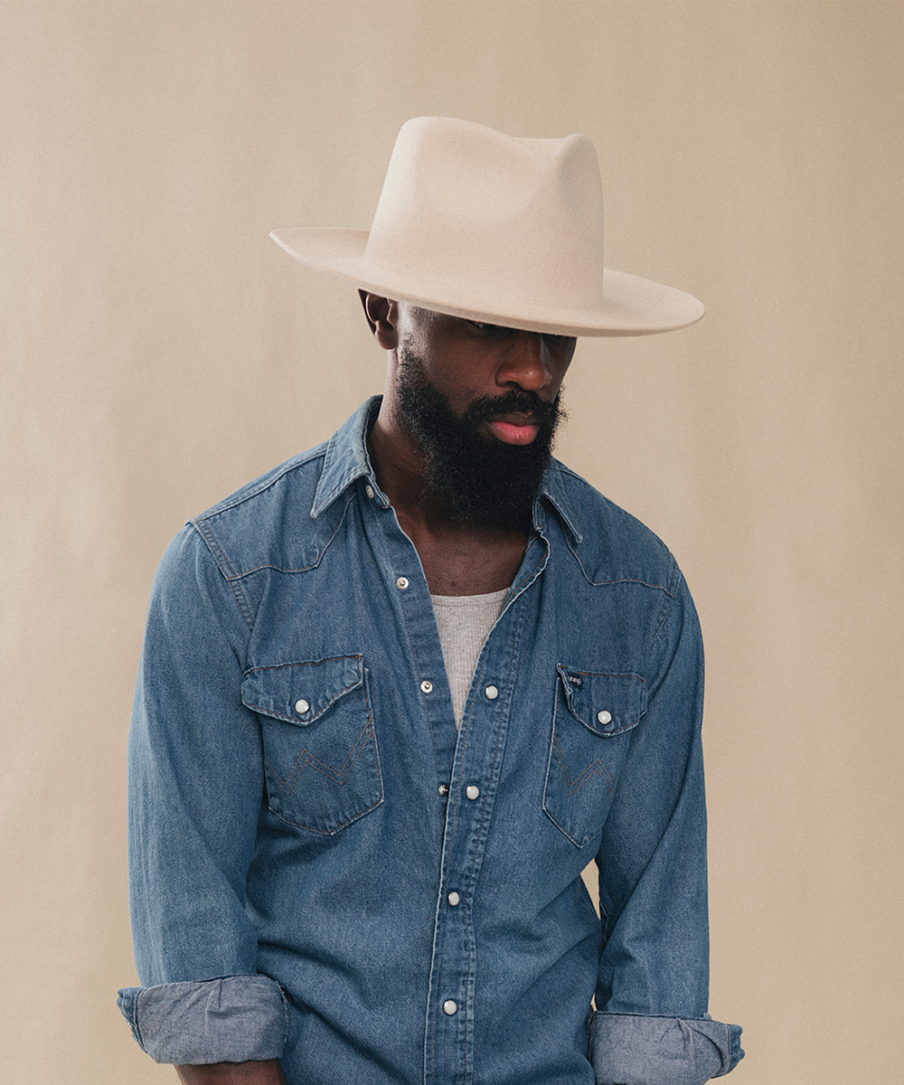 A man wearing a light beige wide-brim fedora hat and a blue denim western-style shirt over a light gray undershirt, posed against a solid beige background #color_buckskin