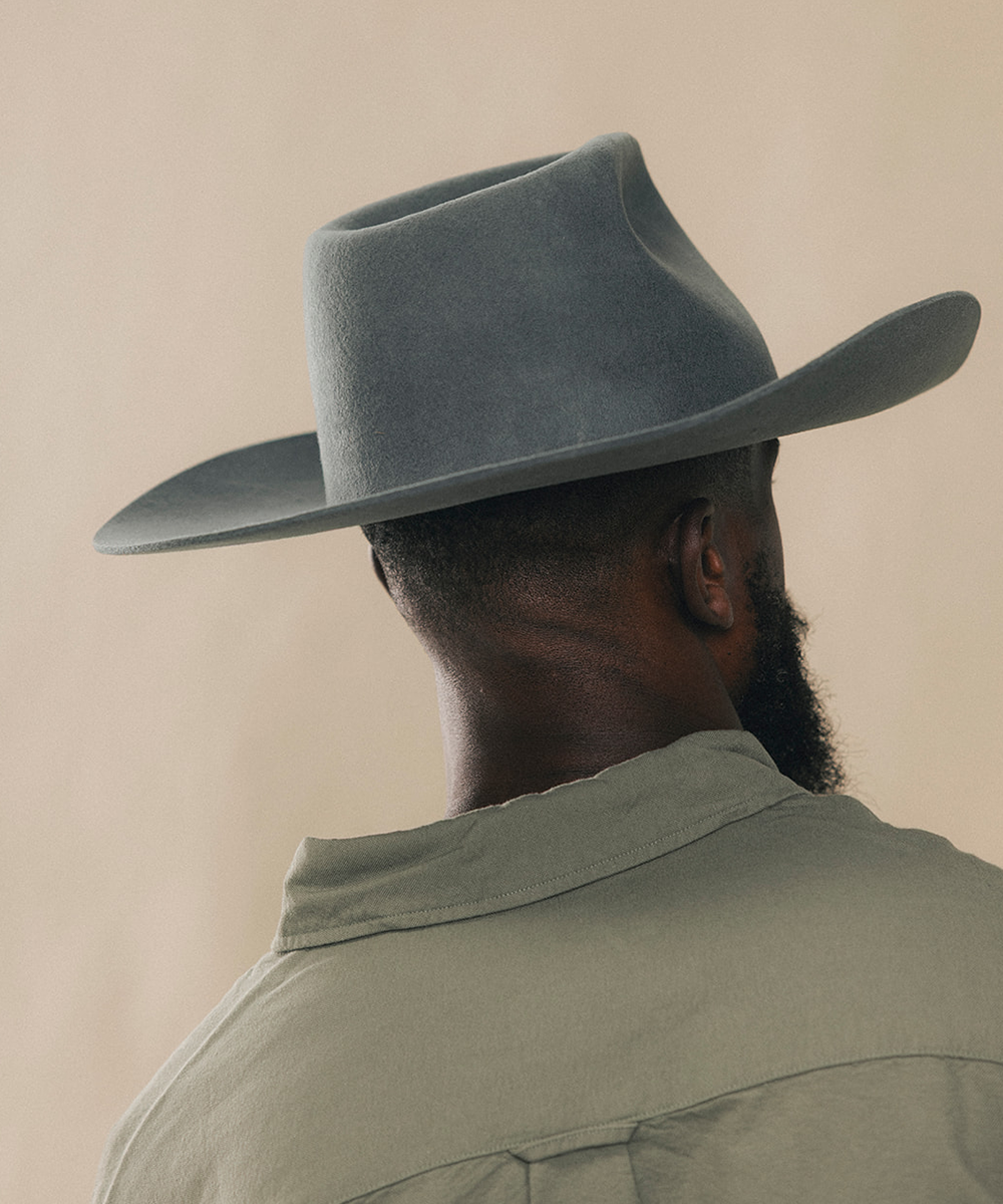A man shown from behind wearing a wide-brimmed felt cowboy hat in a muted blue-gray color, paired with a light olive green shirt against a plain beige background #color_dark grey