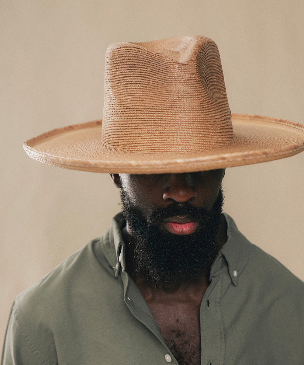 A man wearing a wide-brimmed tan straw hat and an olive green button-down shirt, shown from the chest up against a plain beige background #color_oak