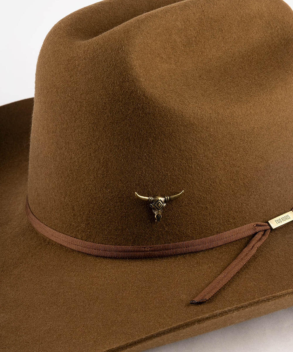 A close-up of a brown felt cowboy hat featuring a matching brown ribbon band with a small gold-toned bull skull pin and a small gold brand tag that reads 'TWO ROADS'. #color_anti gold