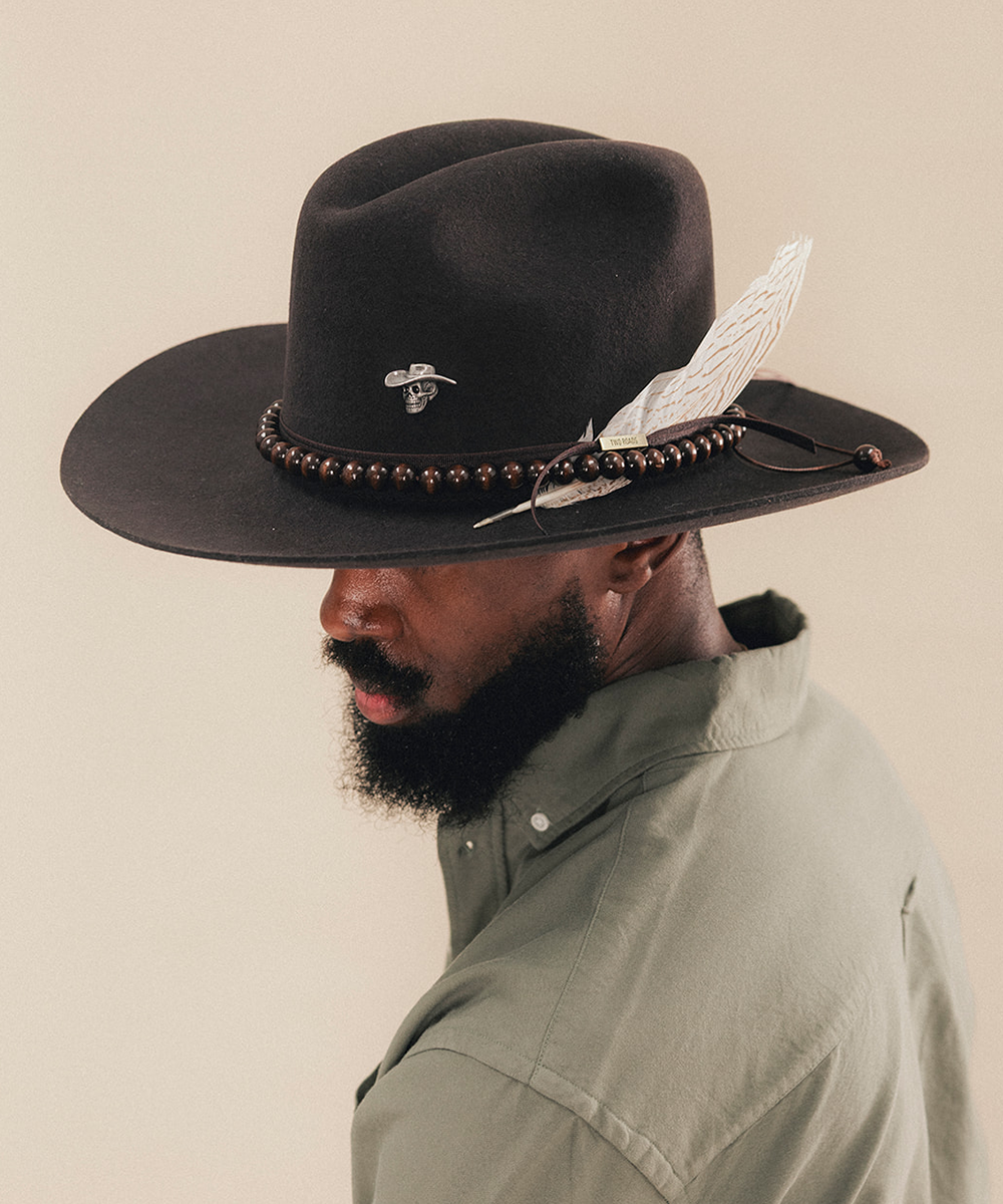 A man in profile wearing a wide-brimmed black cowboy hat with a decorative brown beaded band, a white feather, and a small bee pin, paired with an olive green shirt against a plain beige background #color_hickory