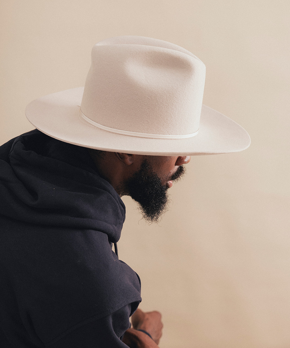 A man wearing a wide-brimmed off-white felt cowboy hat and a dark navy hoodie, shown in profile against a warm beige background #color_silverbelly