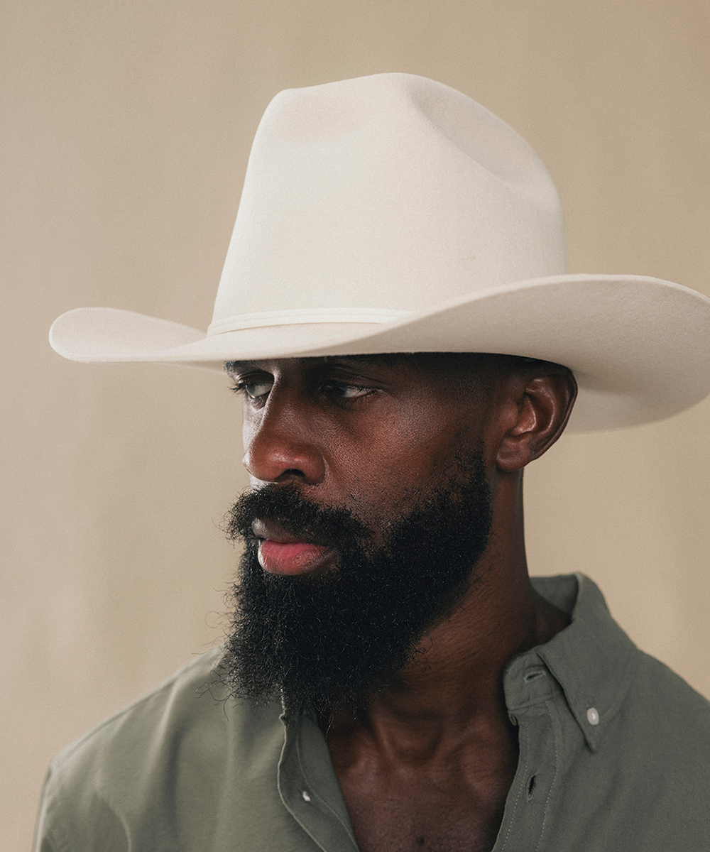 A close-up portrait of a man with a thick black beard wearing a white cowboy hat and an olive green button-down shirt, set against a plain beige background #color_bone