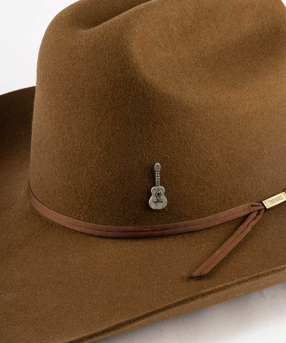 A close-up of a brown felt cowboy hat featuring a matching brown ribbon band with a small silver guitar-shaped pin #color_anti silver