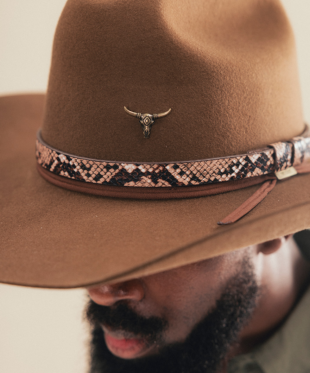 A close-up of a man wearing a brown felt cowboy hat with a patterned band and a small bull skull ornament on the front #color_anti gold