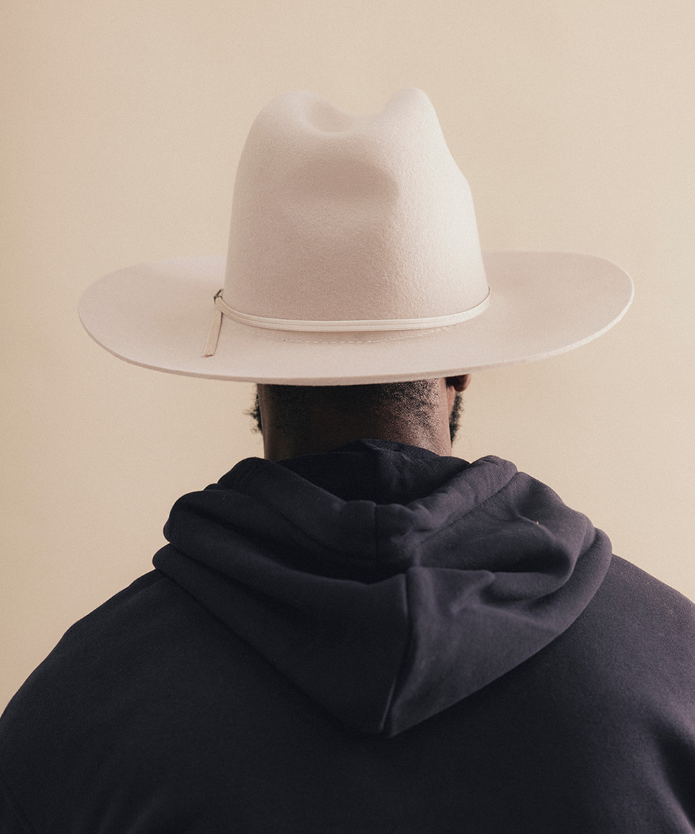 A person shown from behind wearing a light cream-colored wide-brim fedora hat and a dark navy hoodie, set against a solid beige background #color_silverbelly
