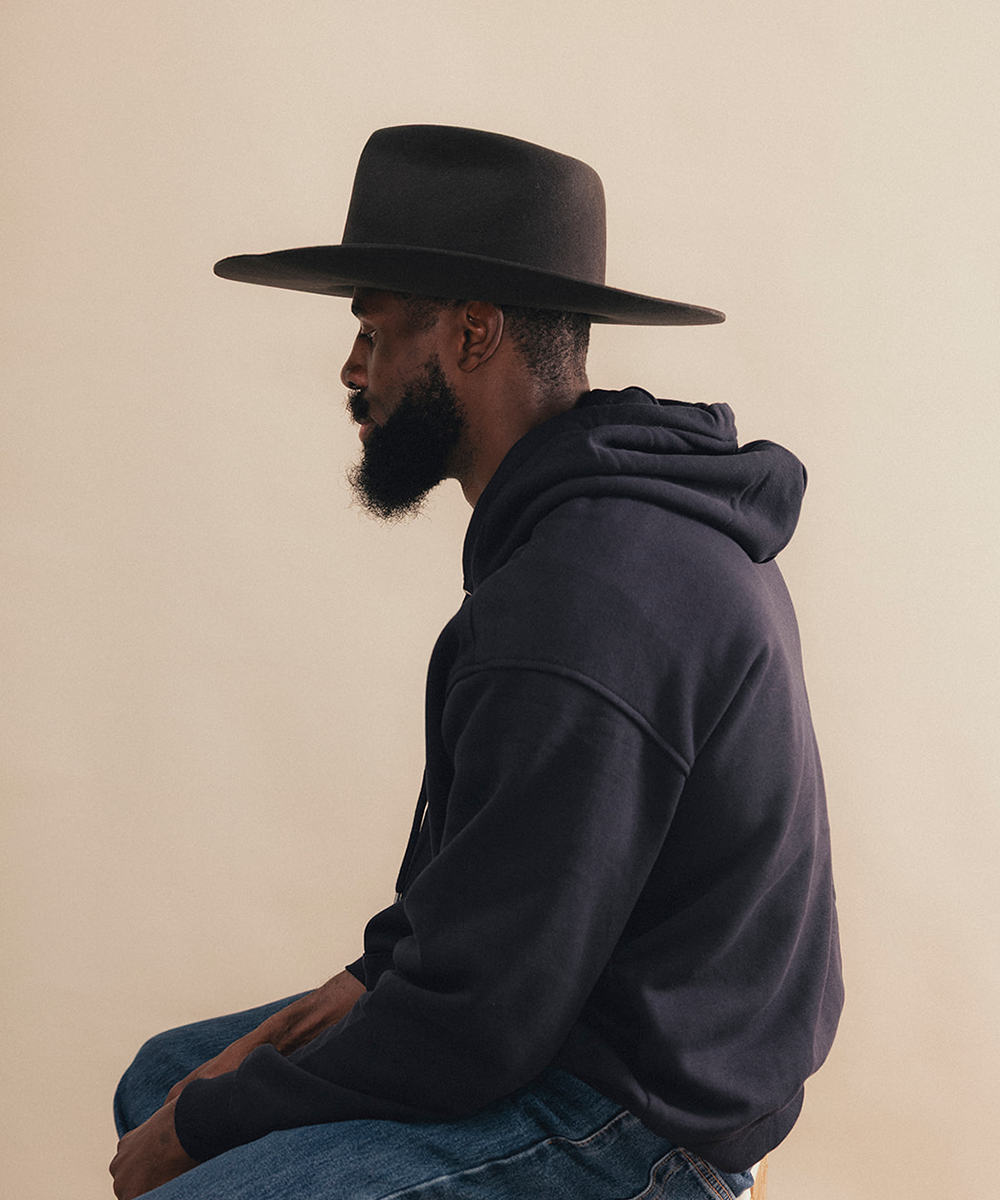 A man in profile wearing a black wide-brim fedora hat, a dark charcoal hoodie, and blue jeans, seated against a solid beige background #color_hickory