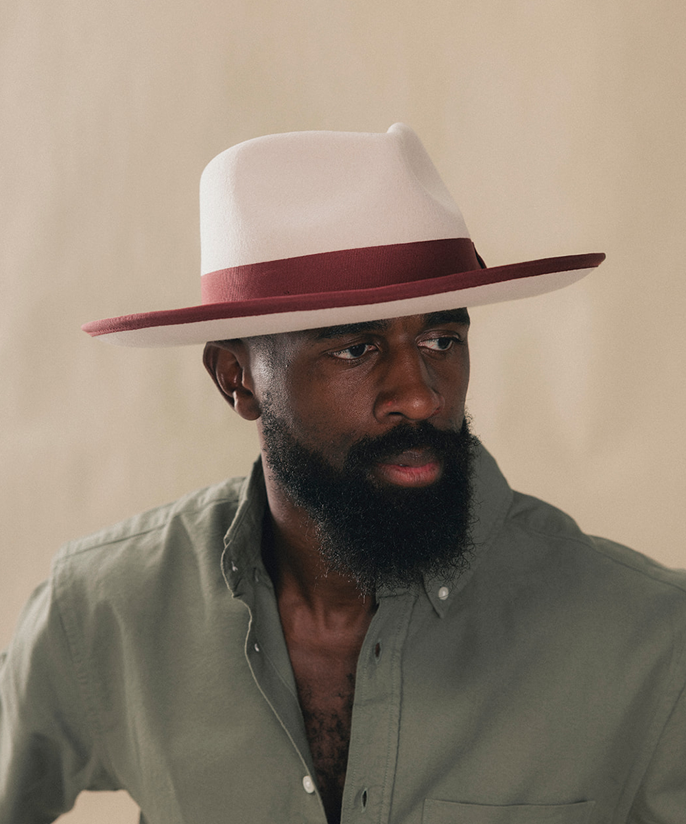 A man with a full beard wearing a white fedora hat with a dark red band and a sage green button-down shirt, photographed against a plain beige background #color_maroon