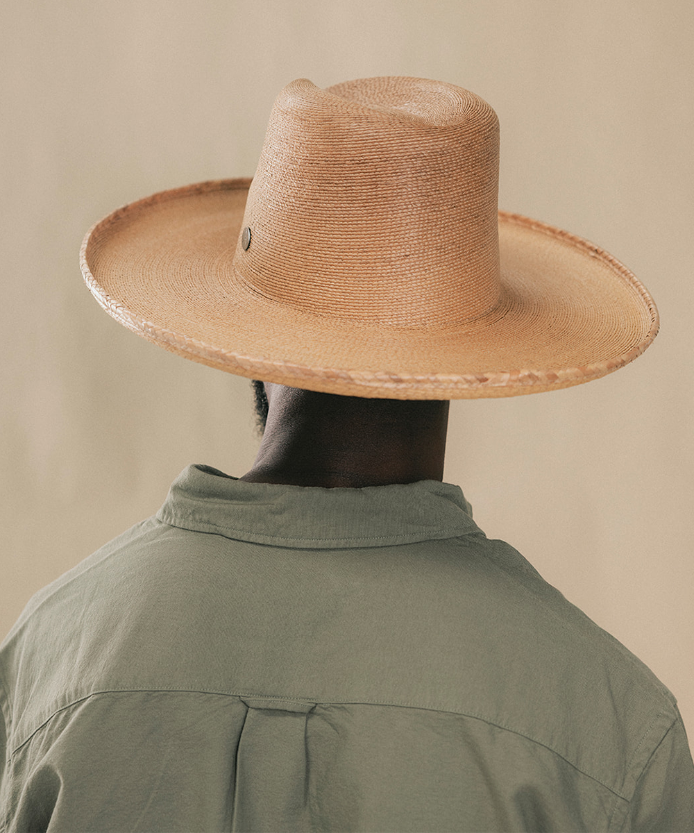 A person shown from the back wearing a wide-brimmed natural straw cowboy hat and an olive green shirt, set against a solid beige background #color_oak
