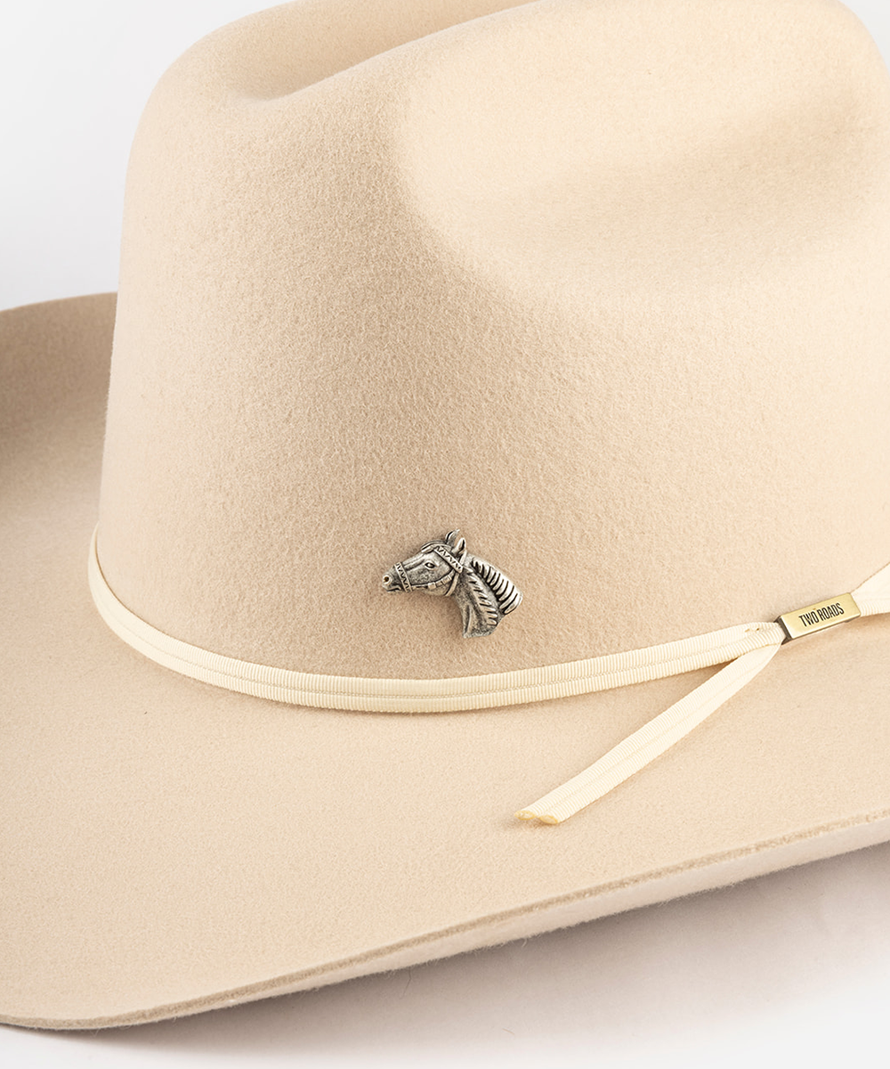 A close-up of a beige felt cowboy hat featuring a small silver horse-shaped pin on the band and a cream-colored ribbon with a gold-toned tip #color_anti silver