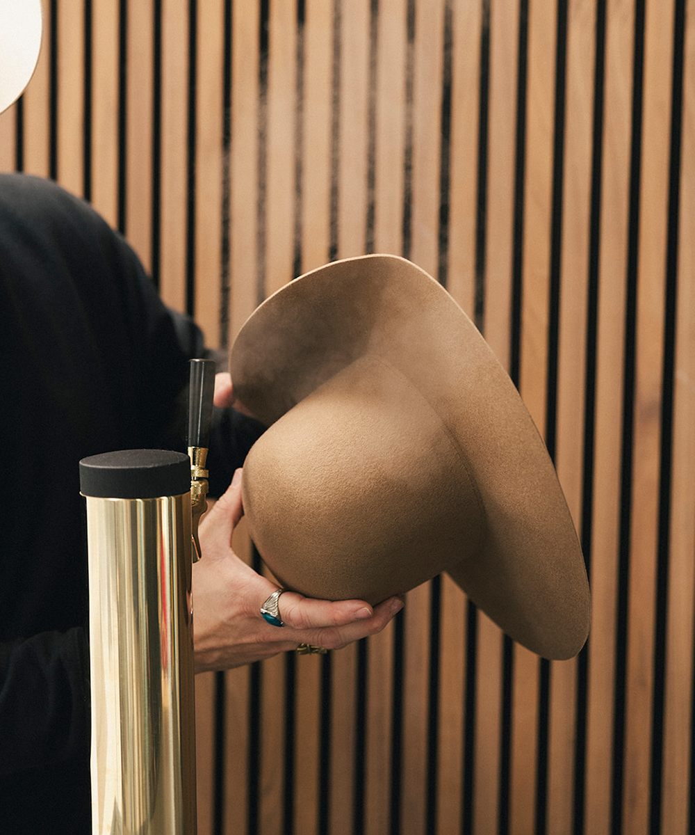 A person in a black outfit holds a wide-brimmed tan hat in front of a vertical wood slat wall, with a gold metal pole visible at the lower left #color_brown