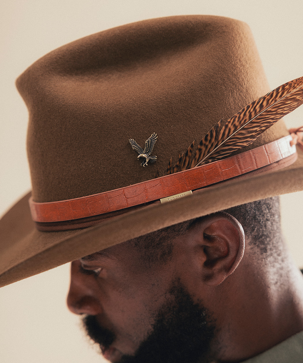 A close-up side profile of a man wearing a brown felt cowboy hat with a reddish-brown band, a feather accent, and a small bird-shaped pin #color_brown