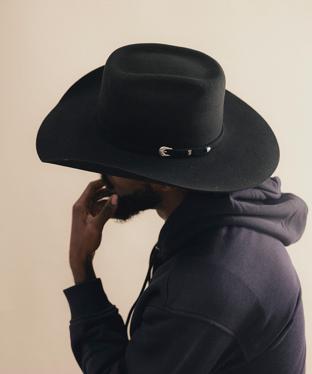 A man in profile wearing a black wide-brim fedora hat with a small silver buckle detail and a dark hoodie, set against a plain beige background #color_black
