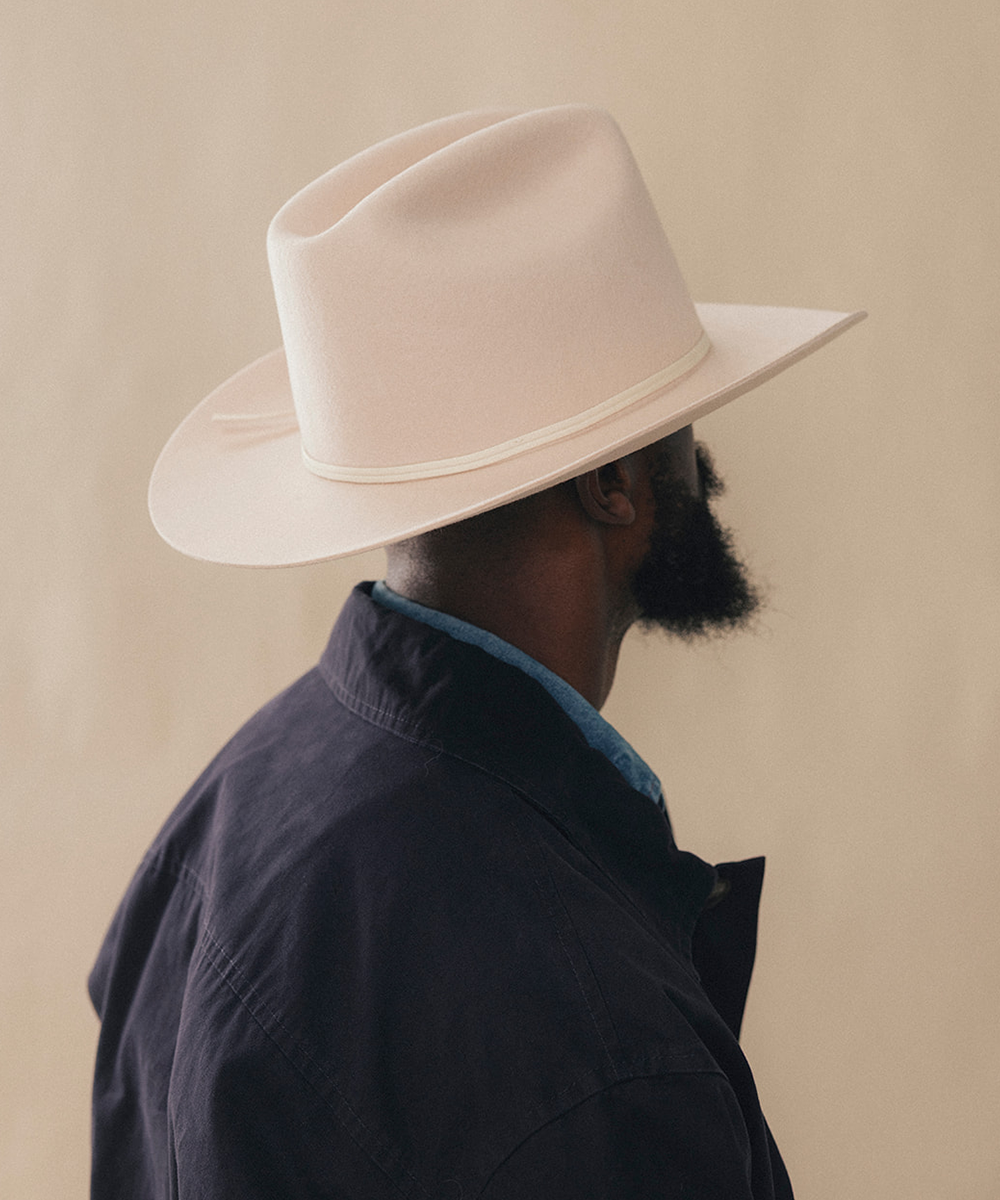 A man shown from the side and back wearing a cream-colored wide-brim fedora hat, a dark navy jacket, and a light blue shirt collar, set against a solid beige background #color_buckskin