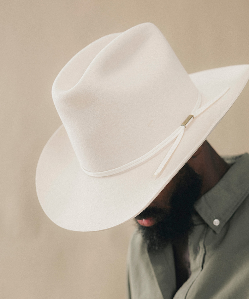A close-up of a man wearing a white felt cowboy hat with a matching white ribbon and a small gold accent, paired with a muted green shirt against a soft beige background #color_bone