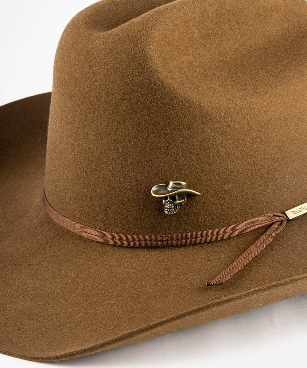 A close-up of a brown felt cowboy hat featuring a matching brown ribbon band with a small metallic skull and hat-shaped ornament, and a small gold brand tag on the side #color_anti gold