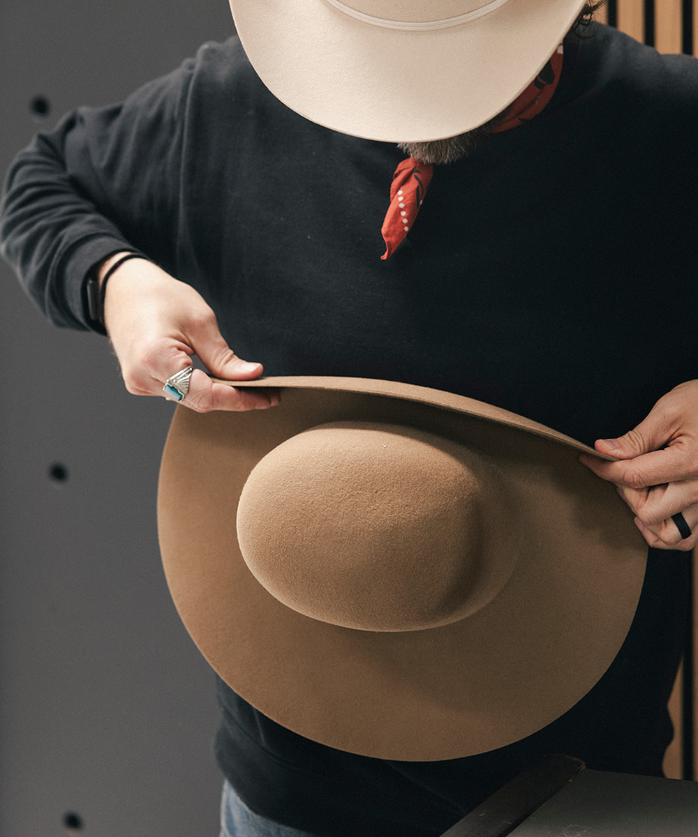 A person wearing a black long-sleeve shirt and a cream cowboy hat holds up a large tan felt wide-brim hat to show its shape and texture #color_brown