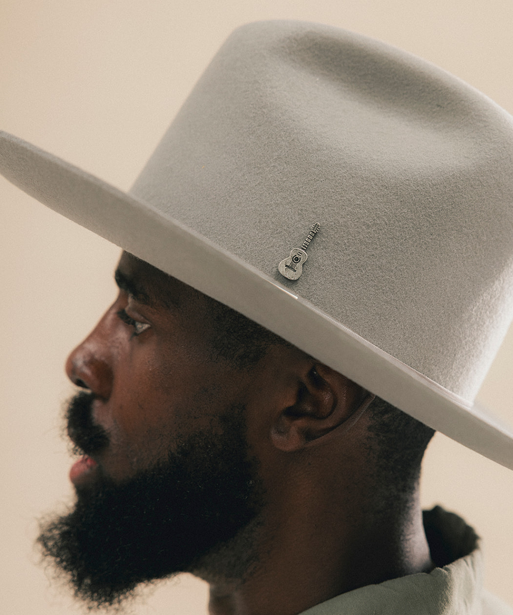 A close-up side profile of a man wearing a light gray felt cowboy hat with a small silver guitar-shaped pin on the side, against a plain beige background #color_anti silver