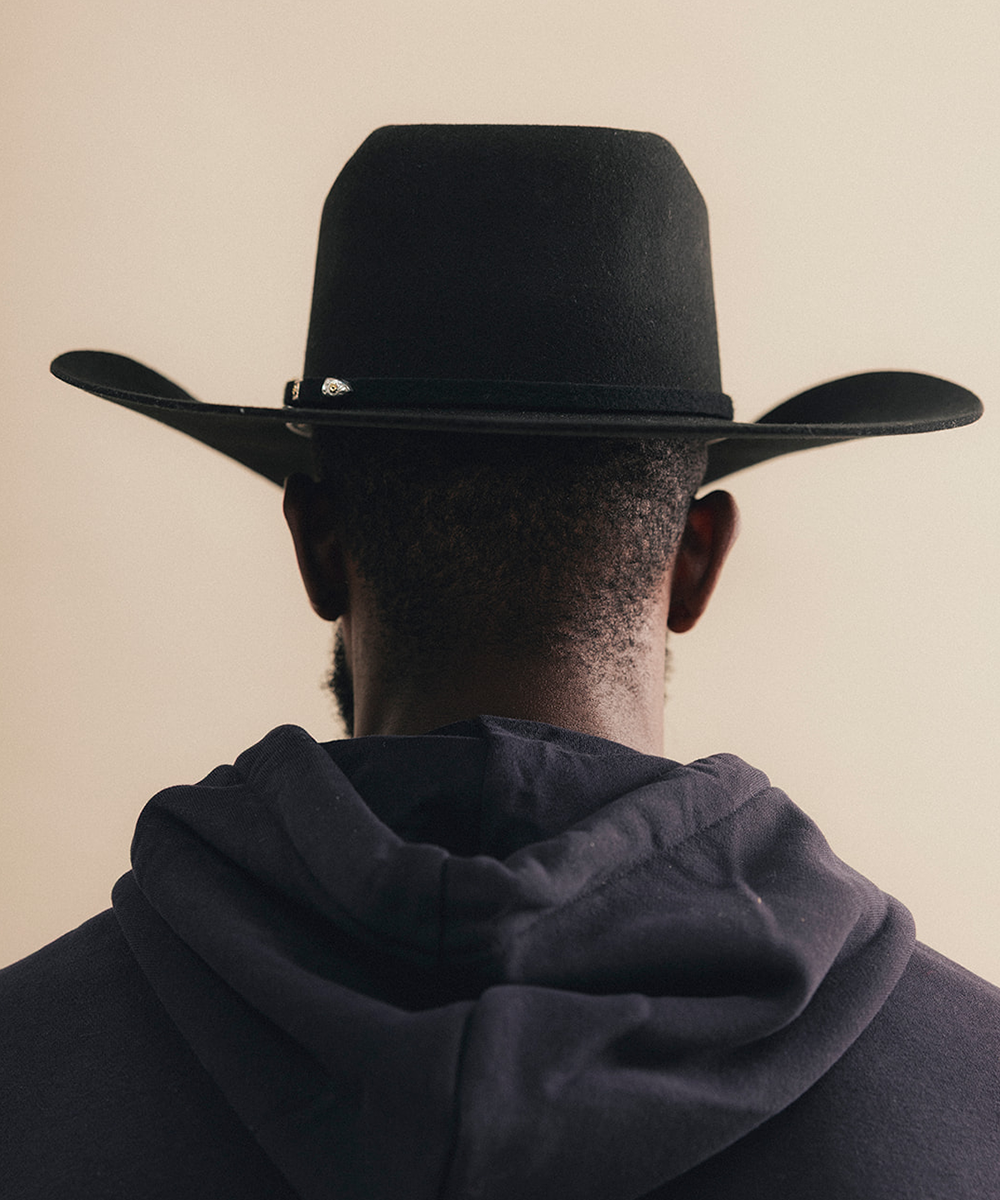 A man viewed from behind wearing a black cowboy hat and a dark hoodie against a plain beige background #color_black