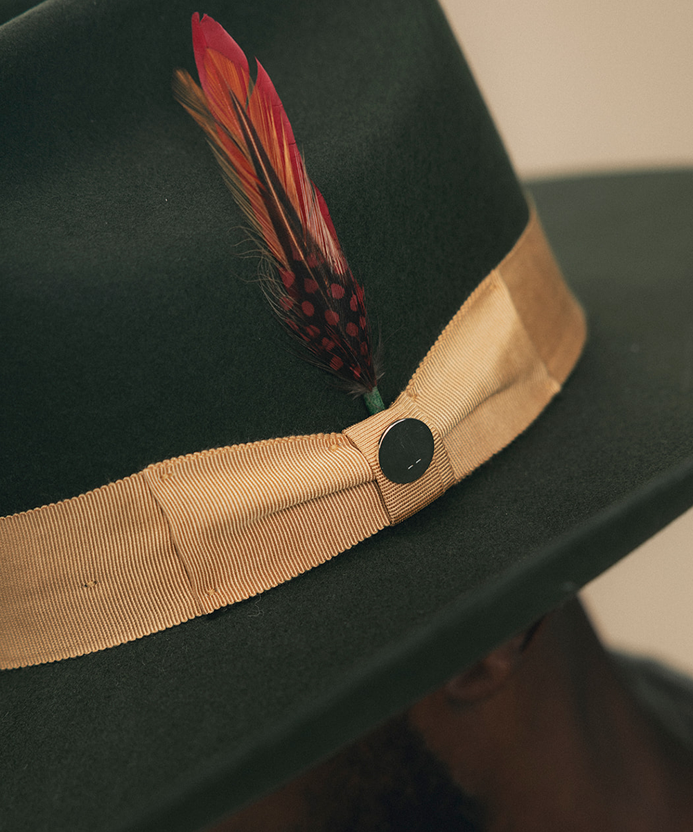 A close-up of a dark green felt fedora hat with a wide tan ribbon band and a decorative red feather accent, worn by a person whose ear and neck are partially visible #color_forest green/mustard
