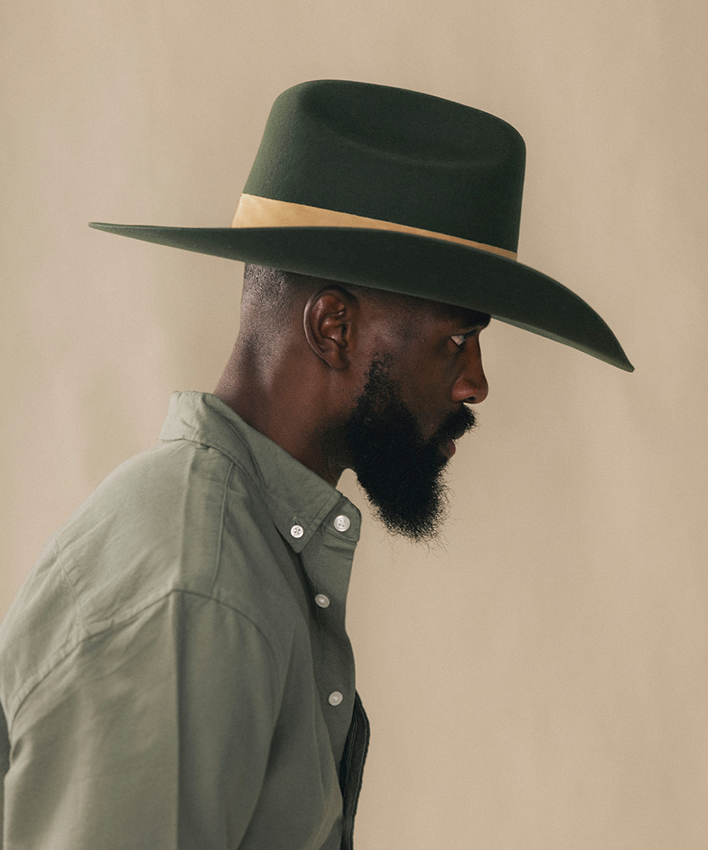 A man in profile wearing a dark green wide-brim cowboy hat with a tan band and an olive green button-down shirt, set against a plain beige background #color_forest green/mustard