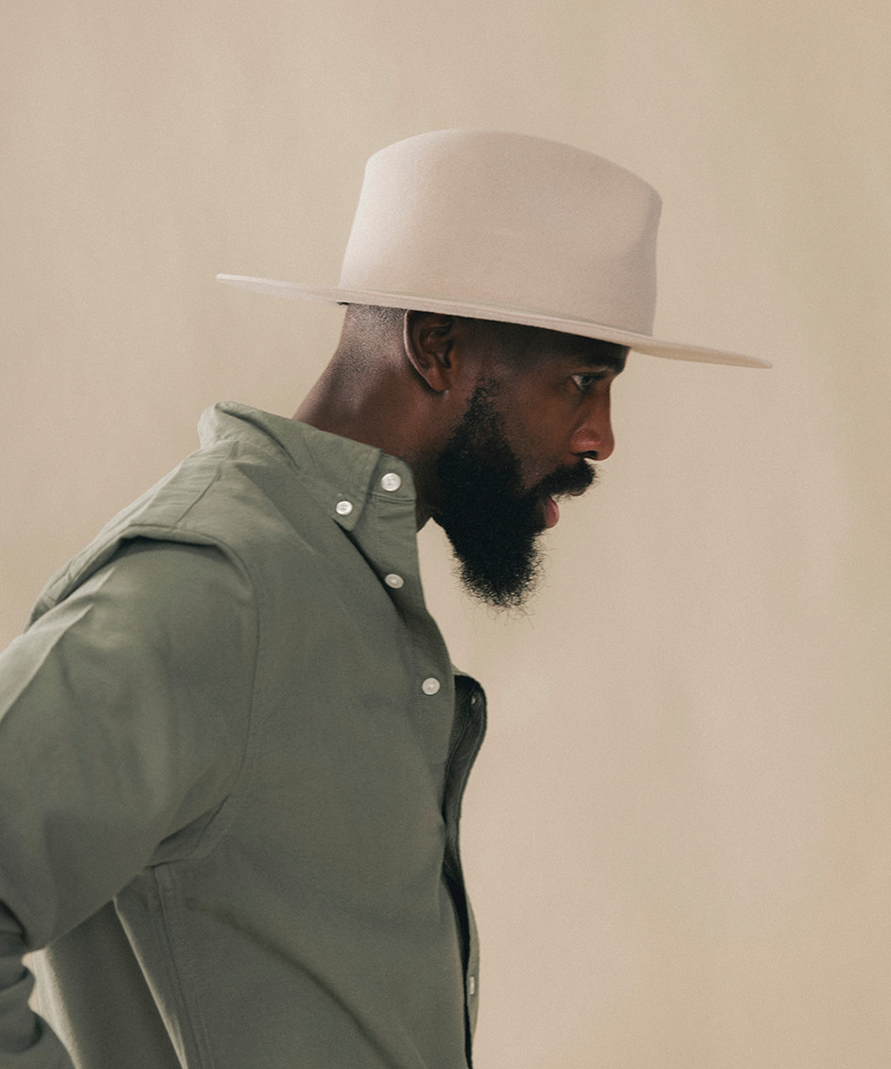 A man in profile wearing a light beige wide-brim fedora hat and an olive green button-down shirt, set against a solid warm beige background #color_silverbelly
