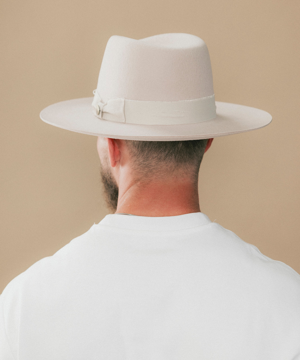 A man wearing a cream felt fedora hat and a white t-shirt, viewed behind against a solid beige background #color_ivory