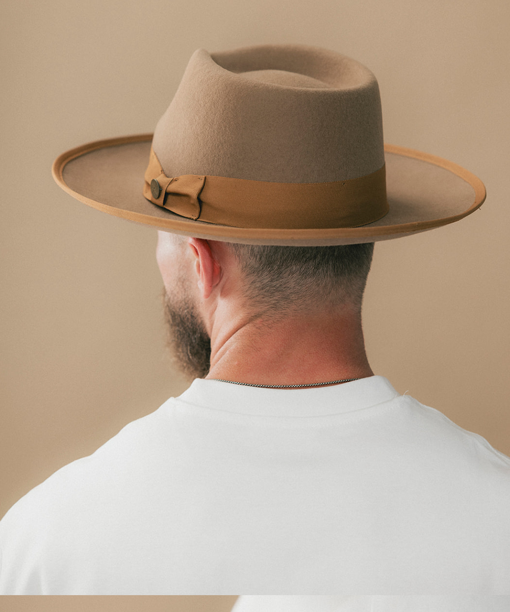 A man shown from the back wearing a wide-brim fedora hat in a warm taupe-brown color with a matching brown band, paired with a white t-shirt against a solid beige background #color_brown