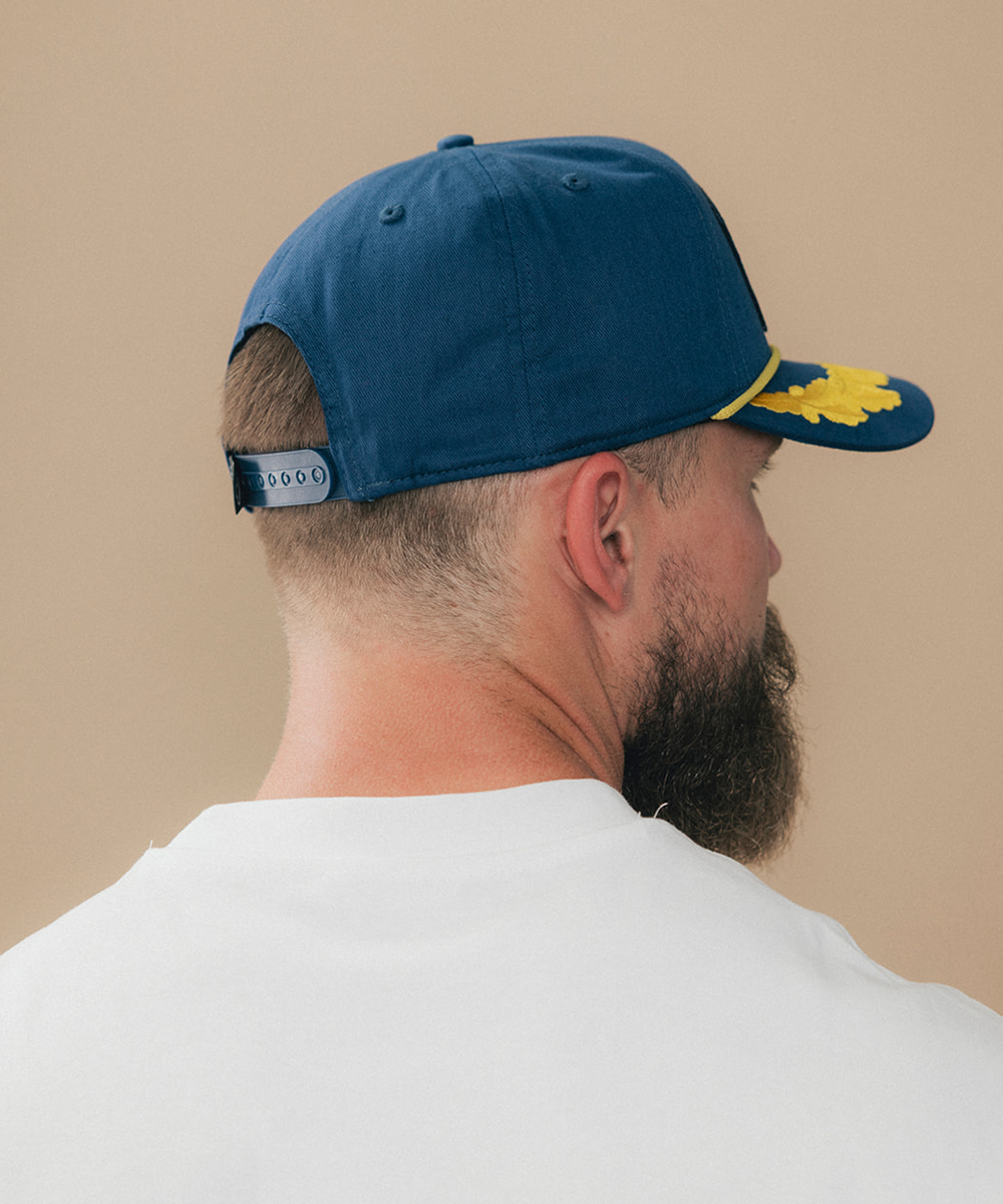 A man shown from the back wearing a navy blue baseball cap with a yellow embroidered logo on the brim and a black adjustable strap #color_blue