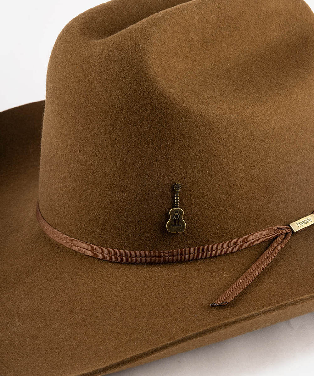 A close-up of a brown felt cowboy hat featuring a matching brown ribbon band with a small guitar-shaped metal pin and a gold-tone brand tag #color_anti gold