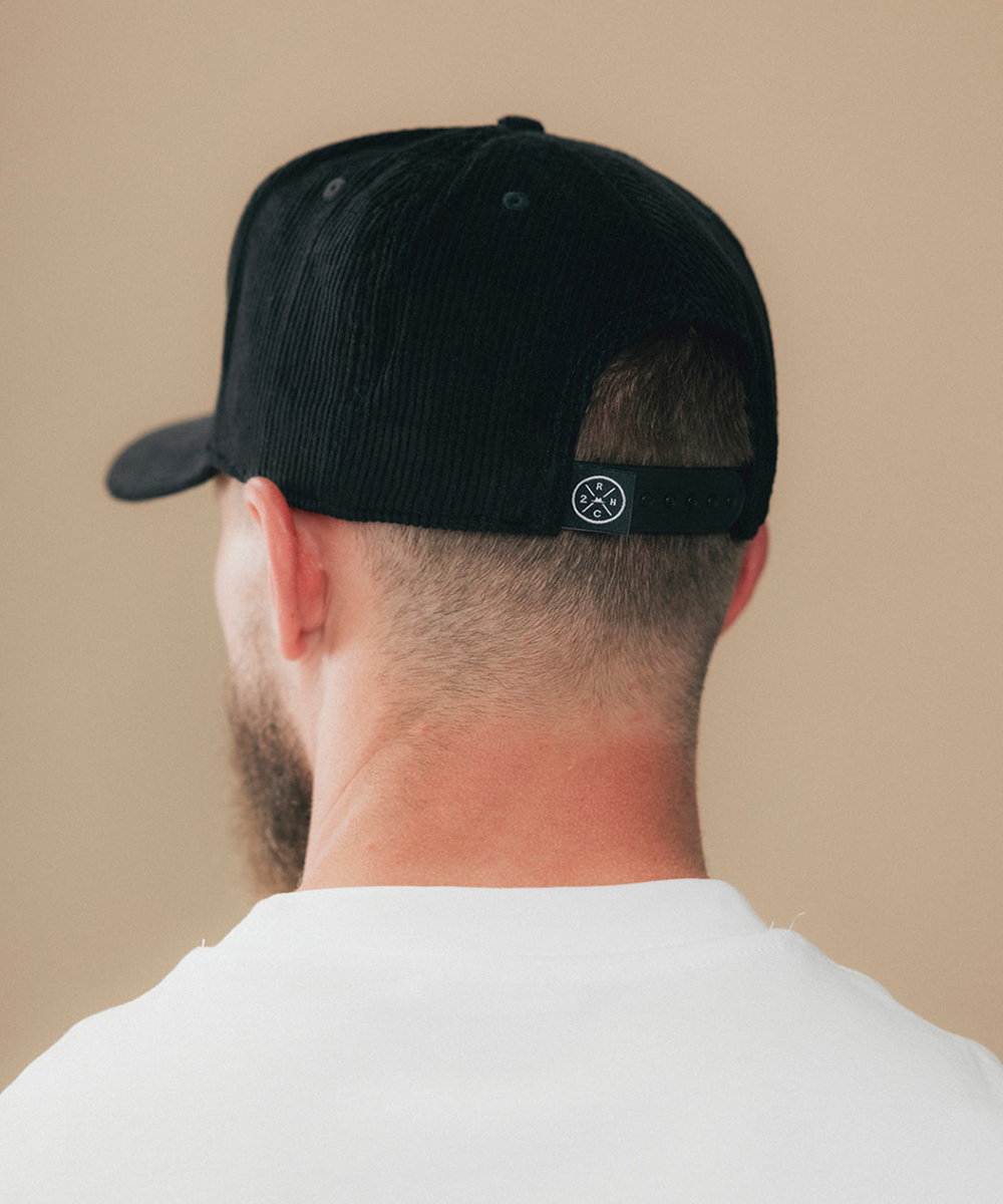 Rear view of a man wearing a black corduroy baseball cap with a black adjustable strap and a small circular logo patch, against a plain beige background #color_obsidian