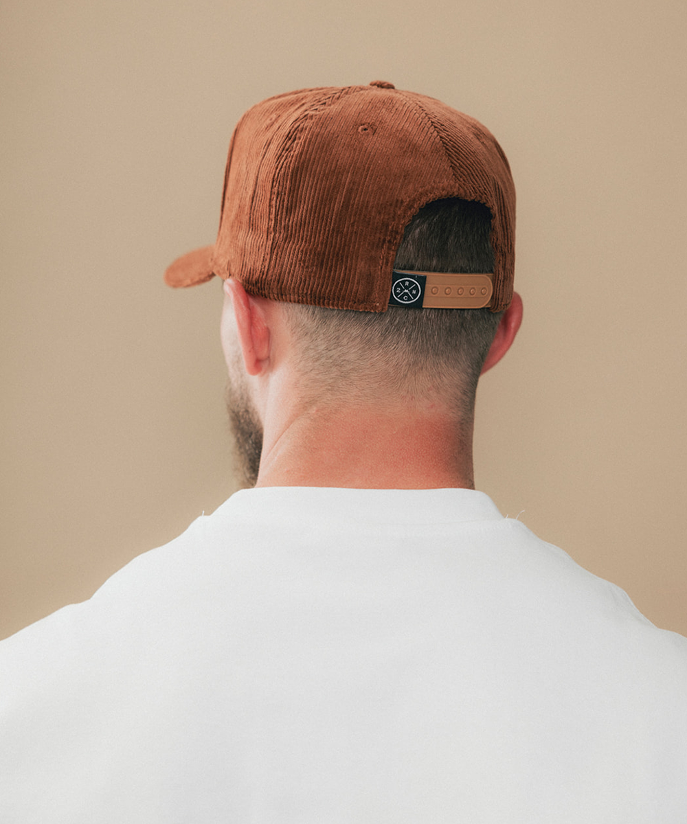 A man viewed from behind wearing a brown corduroy baseball cap with a tan leather brand patch on the back, paired with a white t-shirt against a solid beige background #color_sienna