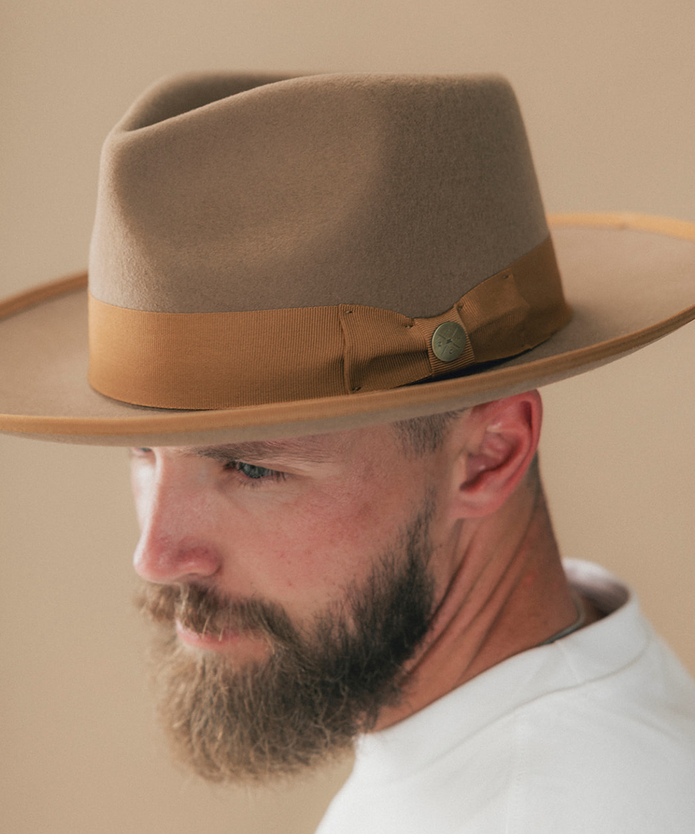 A close-up side profile of a man wearing a wide-brim fedora hat. The hat is light tan with a medium brown band and a small round button detail #color_brown