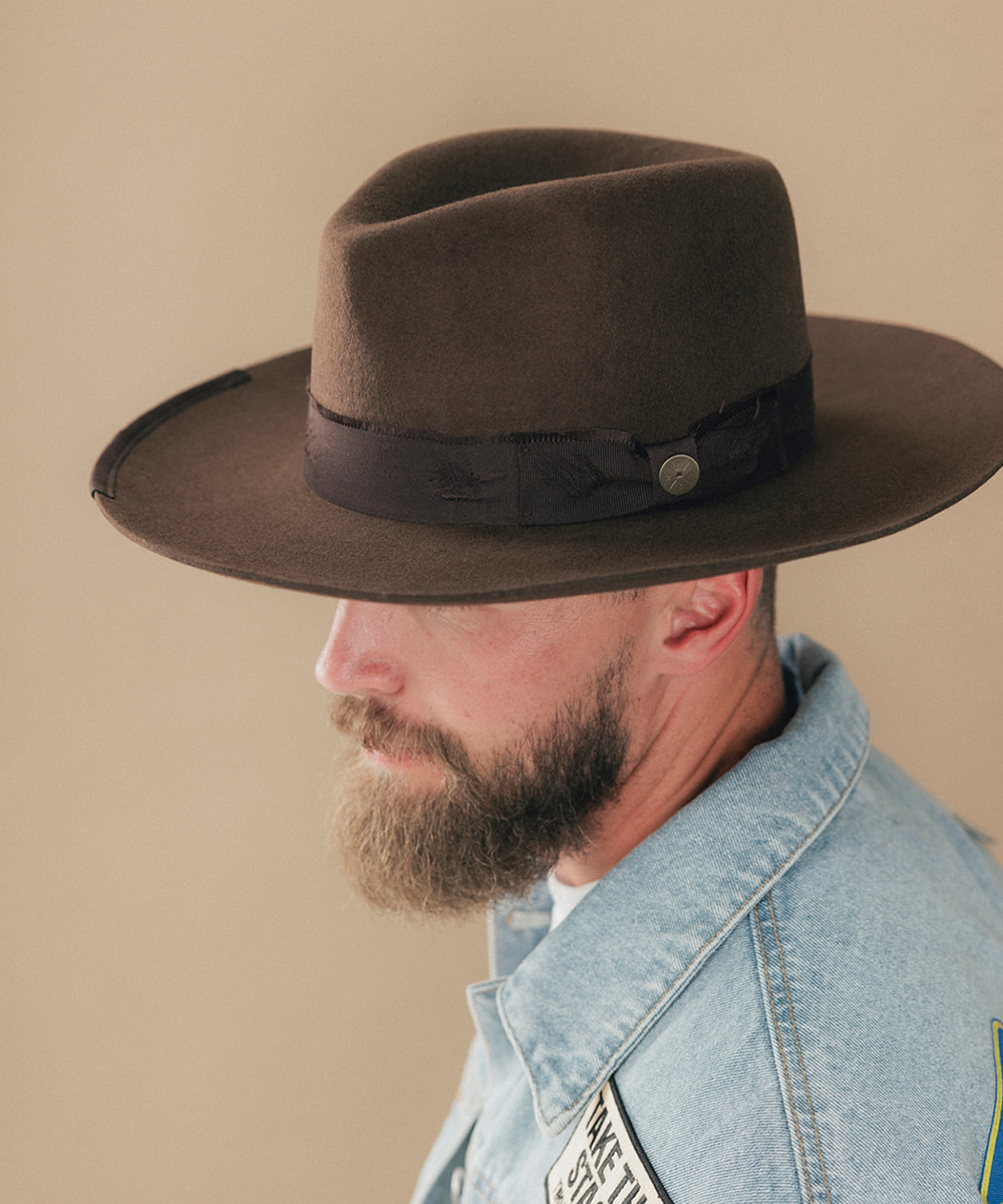 A man with a full beard wearing a dark brown felt fedora hat and a denim jacket, shown in a side profile against a solid beige background #color_dark brown