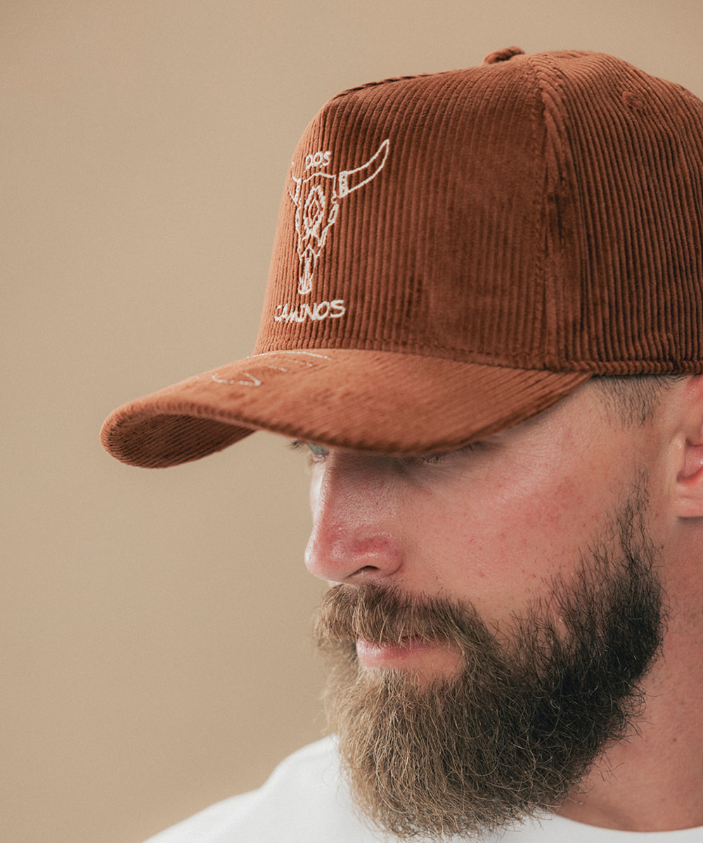 A close-up side profile of a man wearing a brown corduroy baseball cap with a white embroidered longhorn logo and the word 'DOS CAMINOS' on the front, against a plain beige background #color_sienna