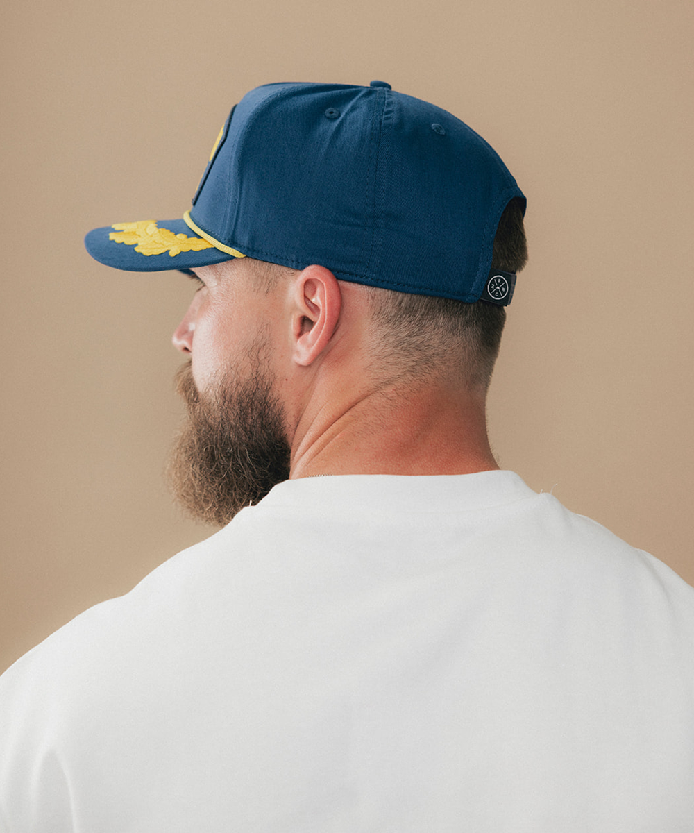 A man shown from the back wearing a navy blue baseball cap with yellow embroidered details on the brim and a small circular logo on the back, paired with a plain white t-shirt against a solid beige background #color_blue