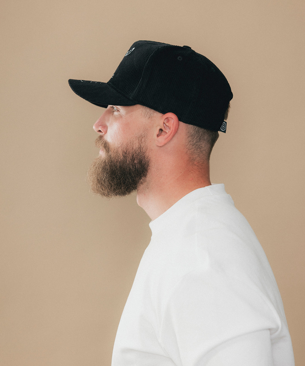 A man in profile wearing a black baseball cap and a white t-shirt, set against a solid beige background #color_obsidian